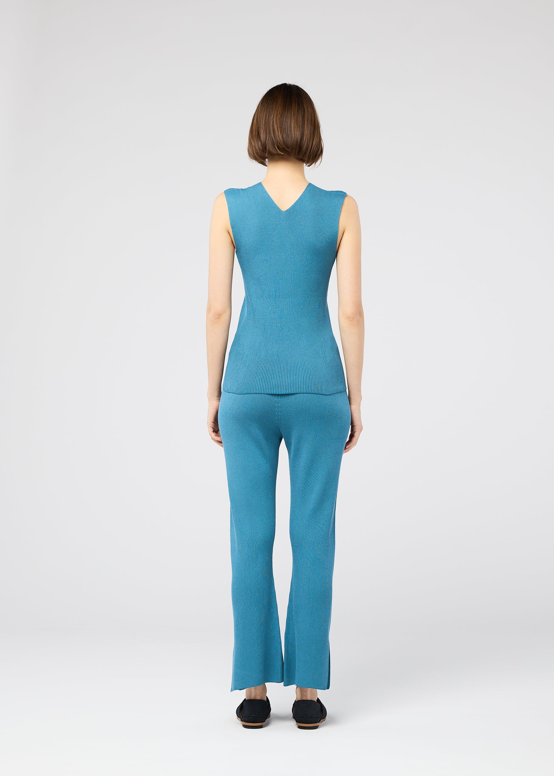 Zoomed view A woman with short brown hair, seen from behind, wears the 132 5. ISSEY MIYAKE FLAT RIB KNIT 2 sleeveless blue jumpsuit and black shoes, standing against a plain light gray background.