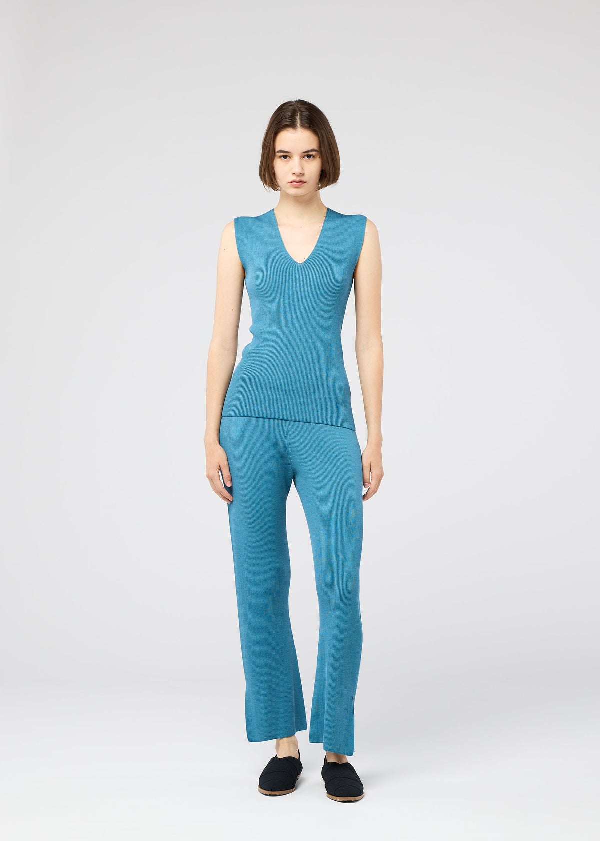 A woman with a bob stands facing forward, wearing the 132 5. ISSEY MIYAKE FLAT RIB KNIT 2 sleeveless blue V-neck top and matching moisture-wicking blue pants, paired with black slip-on shoes against a plain light background. view 9 of 9