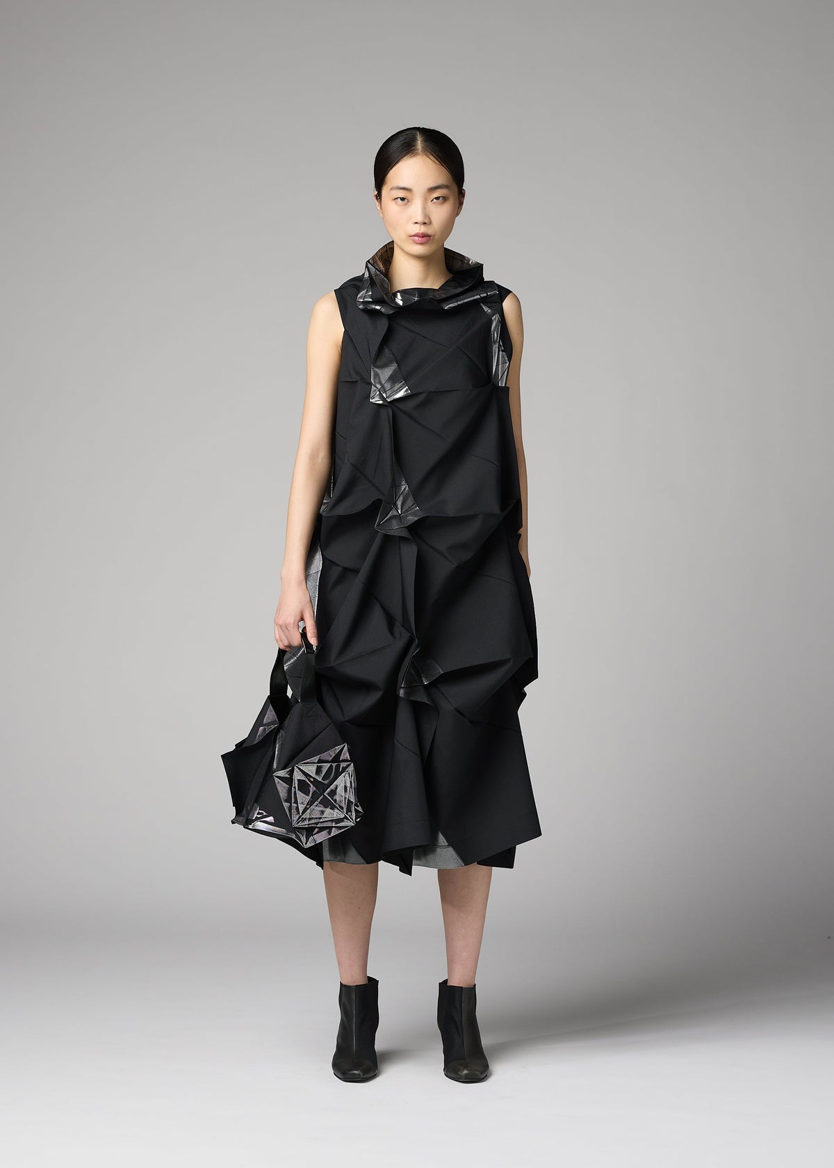 A woman poses against a plain gray background, wearing a sleeveless black dress with geometric folds and holding the No.1-BG bag by 132 5. ISSEY MIYAKE, styled with black ankle boots. view 6 of 7