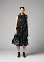 A woman poses against a plain gray background, wearing a sleeveless black dress with geometric folds and holding the No.1-BG bag by 132 5. ISSEY MIYAKE, styled with black ankle boots.