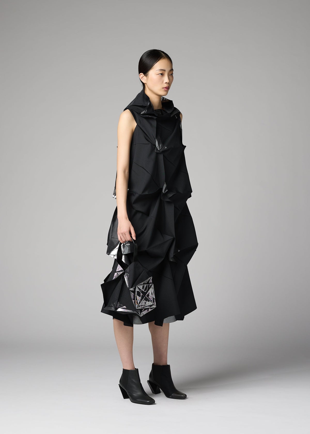 Against a gray backdrop, a woman wears a black sculptural dress with angular folds by 132 5. ISSEY MIYAKE and carries the No.1-BG bag. She pairs the look with black ankle boots, her hair pulled back, and maintains a neutral expression. view 7 of 7