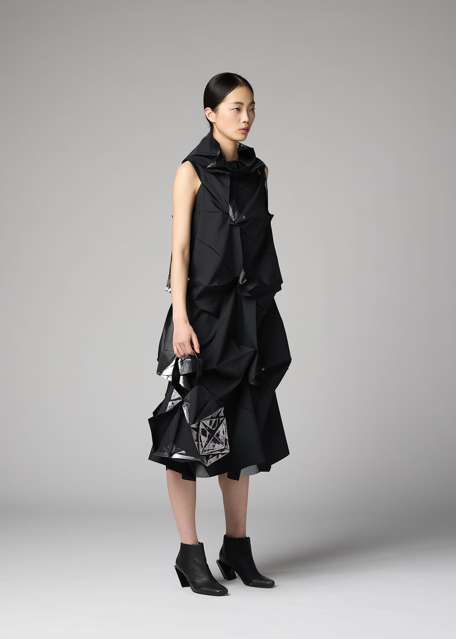 Zoomed view Against a gray backdrop, a woman wears a black sculptural dress with angular folds by 132 5. ISSEY MIYAKE and carries the No.1-BG bag. She pairs the look with black ankle boots, her hair pulled back, and maintains a neutral expression.