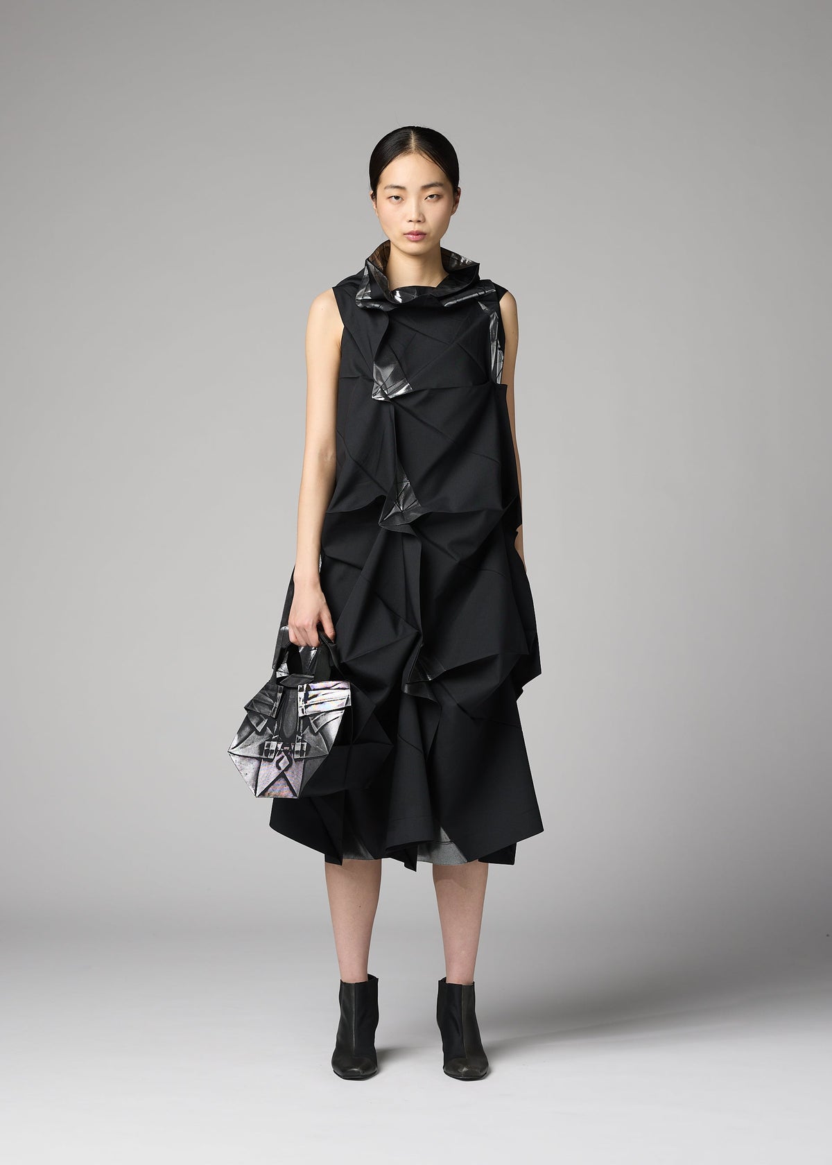 A woman stands against a gray background in the sleeveless, geometric No.6-BG dress by 132 5. ISSEY MIYAKE, holding a matching folding bag and wearing black ankle boots. Her hair is parted in the middle and tied back. view 6 of 6