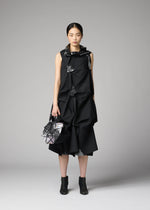 A woman stands against a gray background in the sleeveless, geometric No.6-BG dress by 132 5. ISSEY MIYAKE, holding a matching folding bag and wearing black ankle boots. Her hair is parted in the middle and tied back.