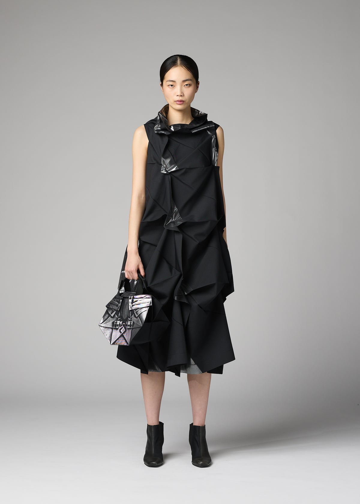 A woman stands against a gray background wearing a black, geometric, textured dress with matching angular handbag and black ankle boots. Her hair is tied back and she has a neutral expression. She carries the 132 5. STANDARD BAG by 132 5. ISSEY MIYAKE—a sleek recycled polyester accessory that seamlessly blends sustainability with sophisticated style. view 6 of 6