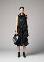 A woman stands against a gray background wearing a black, geometric, textured dress with matching angular handbag and black ankle boots. Her hair is tied back and she has a neutral expression. She carries the 132 5. STANDARD BAG by 132 5. ISSEY MIYAKE—a sleek recycled polyester accessory that seamlessly blends sustainability with sophisticated style.