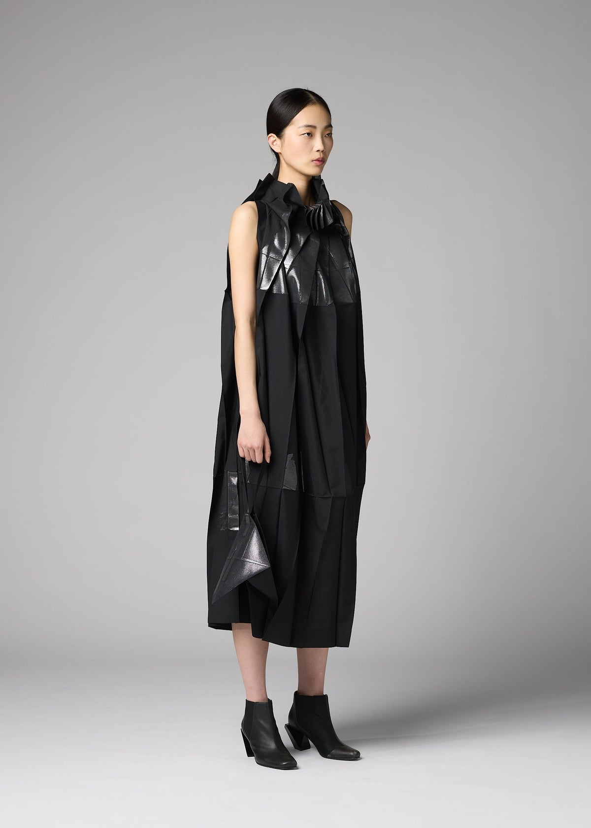 A woman stands in a studio wearing a sleeveless, black, avant-garde dress with shiny and matte textures, asymmetrical details, and black ankle boots. She holds the F-BG bag by 132 5. ISSEY MIYAKE—a sleek foil processed accessory that complements her ensemble. Her straight dark hair is pulled back and she has a neutral expression. view 7 of 7