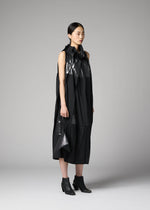 A woman stands in a studio wearing a sleeveless, black, avant-garde dress with shiny and matte textures, asymmetrical details, and black ankle boots. She holds the F-BG bag by 132 5. ISSEY MIYAKE—a sleek foil processed accessory that complements her ensemble. Her straight dark hair is pulled back and she has a neutral expression.