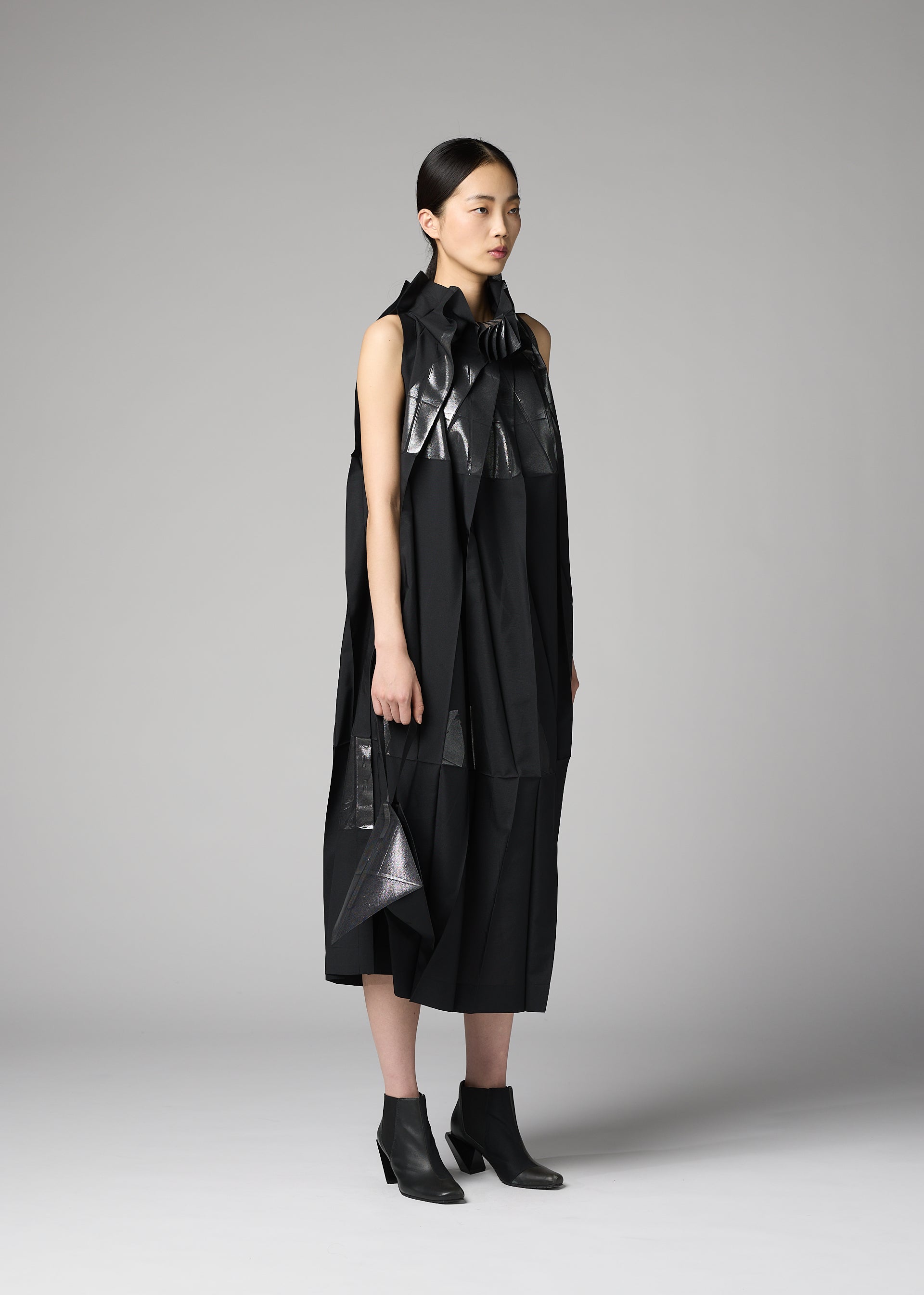 Zoomed view A woman stands in a studio wearing a sleeveless, black, avant-garde dress with shiny and matte textures, asymmetrical details, and black ankle boots. She holds the F-BG bag by 132 5. ISSEY MIYAKE—a sleek foil processed accessory that complements her ensemble. Her straight dark hair is pulled back and she has a neutral expression.