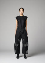 A woman stands facing forward against a gray background, wearing F-BT folding pants from 132 5. ISSEY MIYAKE—wide-leg black with sheer geometric panels—styled with a sleeveless top, black shoes, and straight dark hair pulled back.