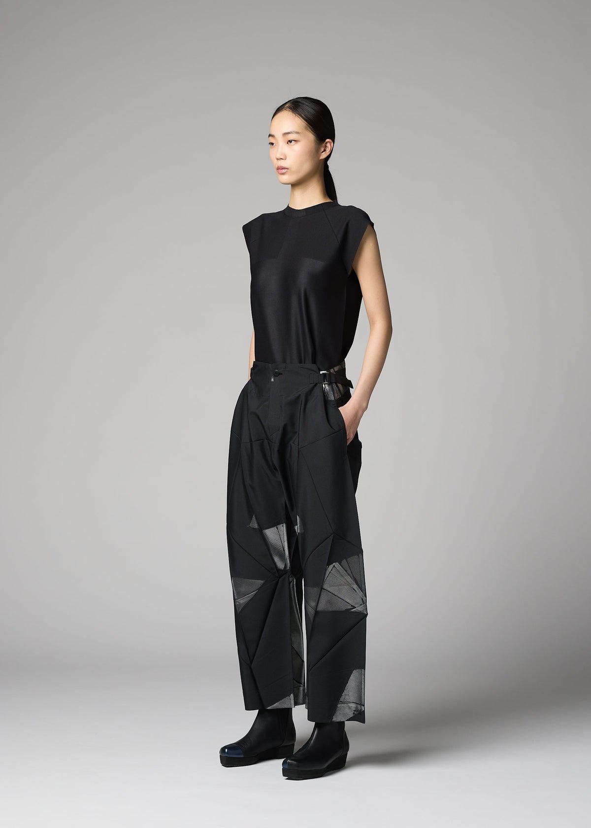 A woman poses against a gray background, wearing 132 5. ISSEY MIYAKE’s F-BT: a sleeveless black top and high-waisted recycled polyester pants with geometric patterns and sheer panels, paired with black boots and hands in her pockets. view 9 of 13