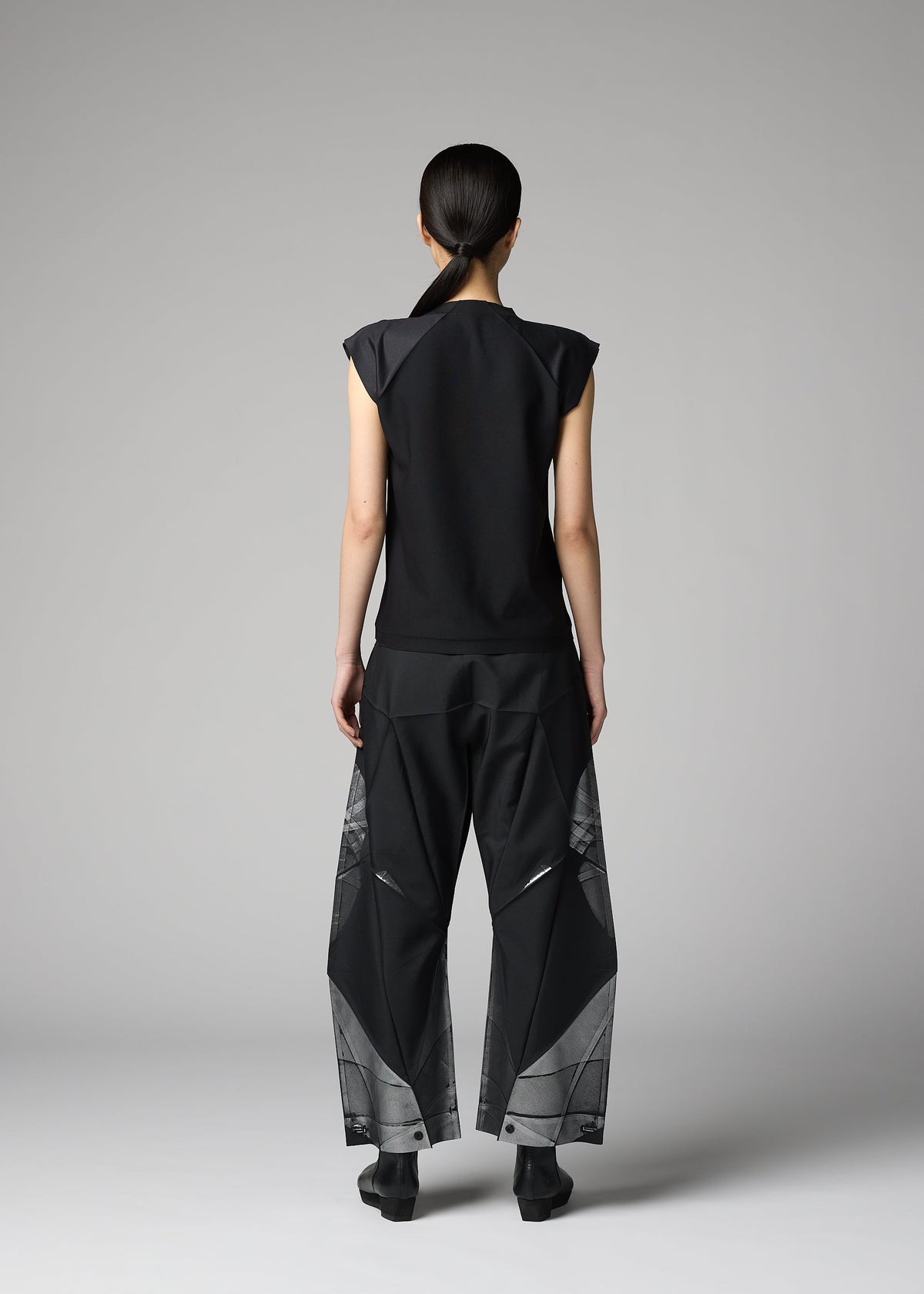 A person with long dark hair in a low ponytail faces away, wearing the 132 5. ISSEY MIYAKE F-BT black sleeveless top and recycled polyester pants with geometric patterns, set against a plain gray background. view 10 of 13