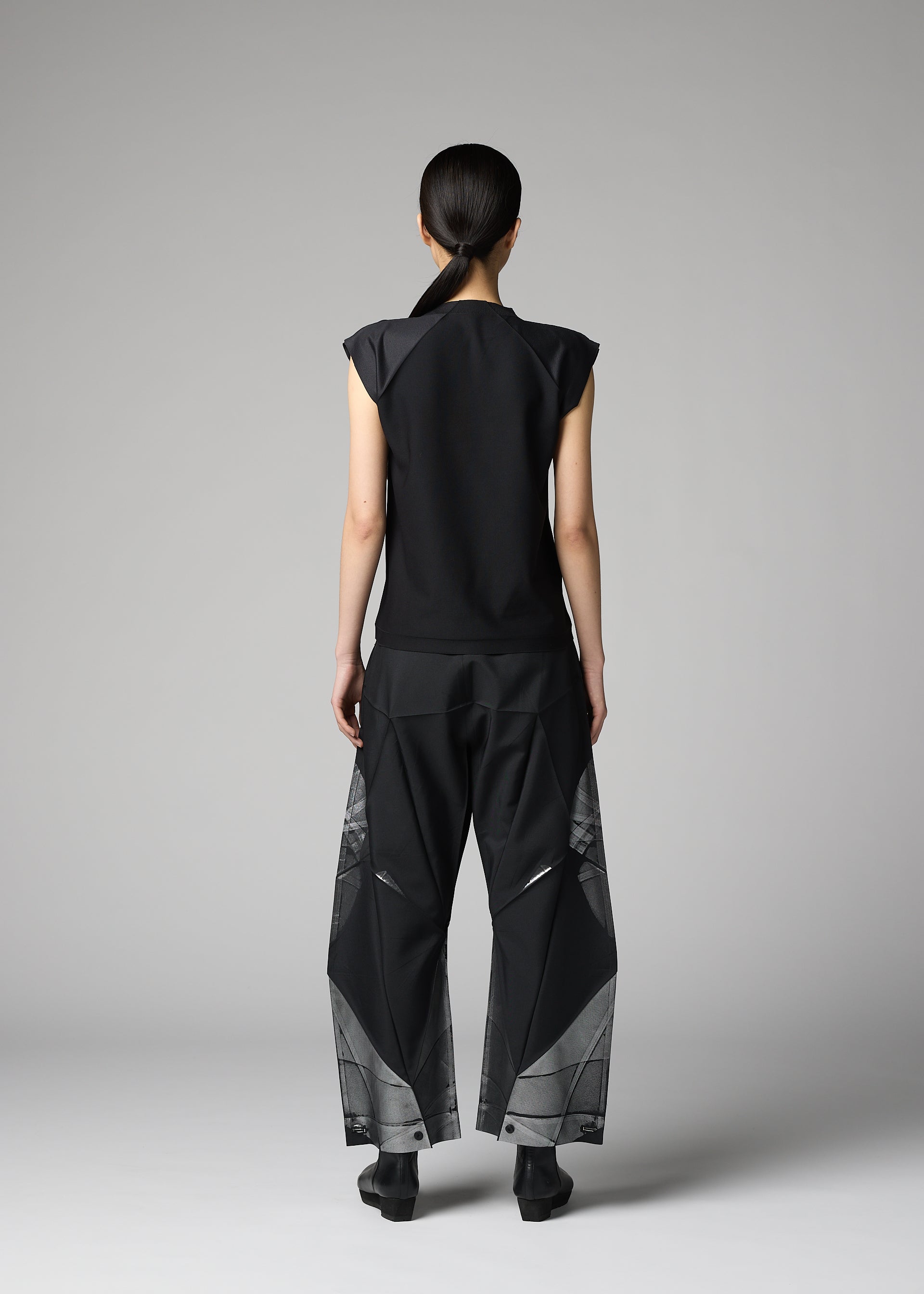 Zoomed view A person with long dark hair in a low ponytail faces away, wearing the 132 5. ISSEY MIYAKE F-BT black sleeveless top and recycled polyester pants with geometric patterns, set against a plain gray background.
