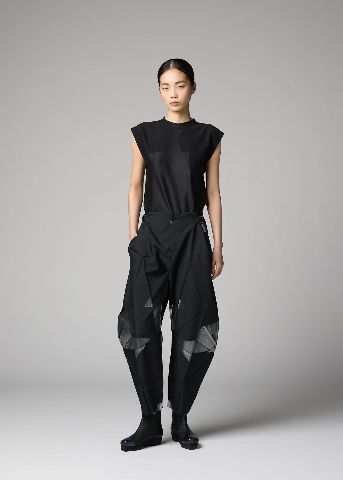 A person stands against a gray backdrop wearing the F-BT black sleeveless top and loose, angular recycled polyester pants with mesh panels by 132 5. ISSEY MIYAKE, paired with black shoes for a modern minimalist look. view 11 of 13
