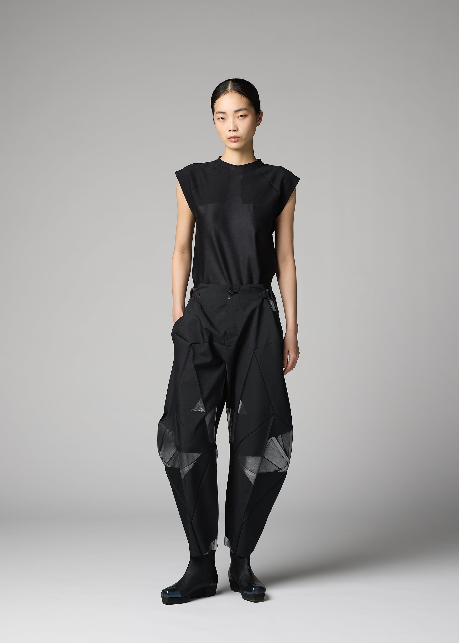 Zoomed view A person stands against a gray backdrop wearing the F-BT black sleeveless top and loose, angular recycled polyester pants with mesh panels by 132 5. ISSEY MIYAKE, paired with black shoes for a modern minimalist look.