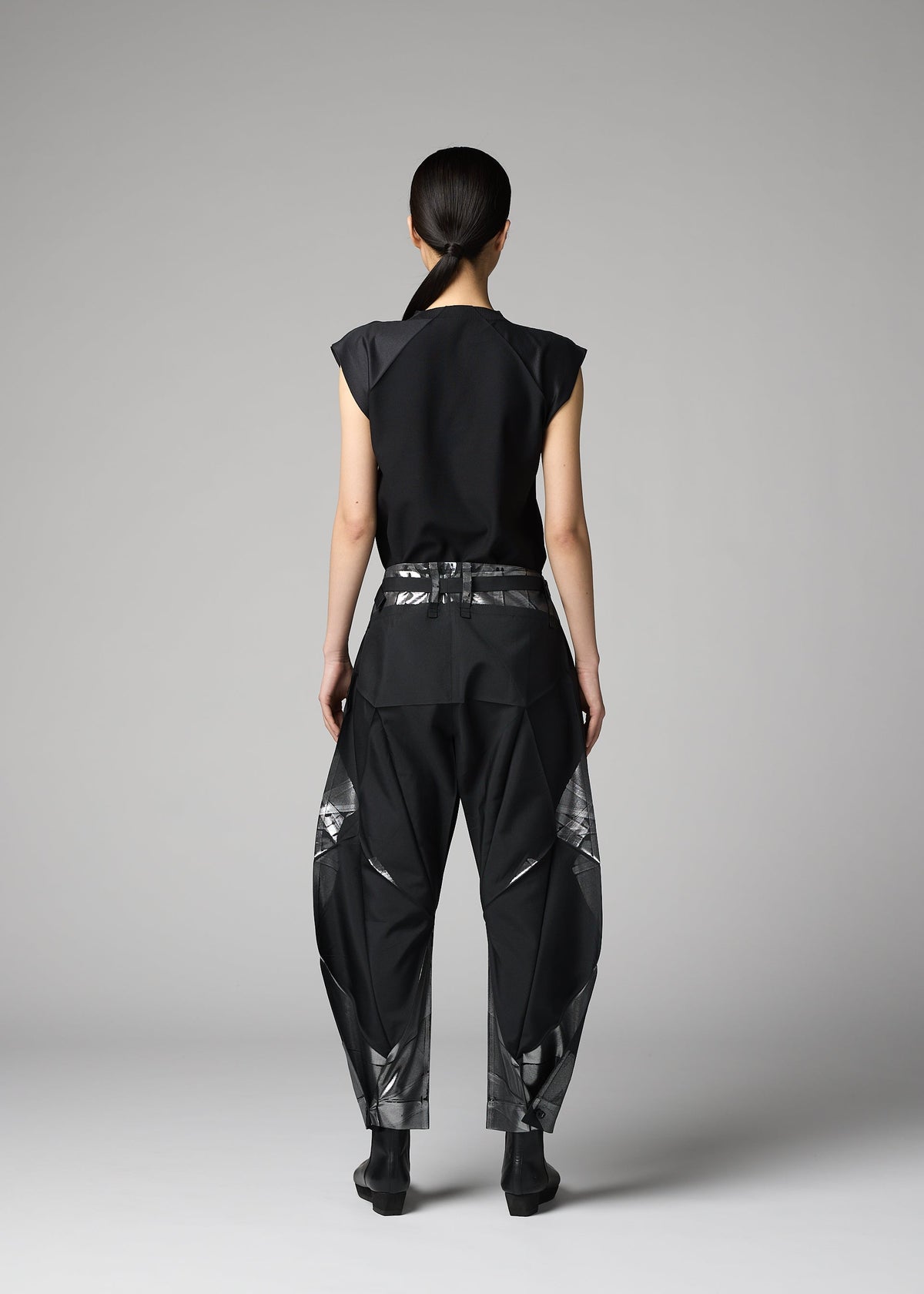 A person with long dark hair tied back faces away, wearing a black sleeveless top and 132 5. ISSEY MIYAKE F-BT folding pants with abstract silver foil details, set against a plain gray background. view 12 of 13
