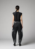 A person with long dark hair tied back faces away, wearing a black sleeveless top and 132 5. ISSEY MIYAKE F-BT folding pants with abstract silver foil details, set against a plain gray background.