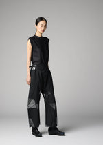 A woman stands in a studio wearing the F-BT from 132 5. ISSEY MIYAKE—sleeveless black top and wide-leg pants with geometric sheer panels, paired with black shoes. She has straight dark hair pulled back, against a plain gray background.