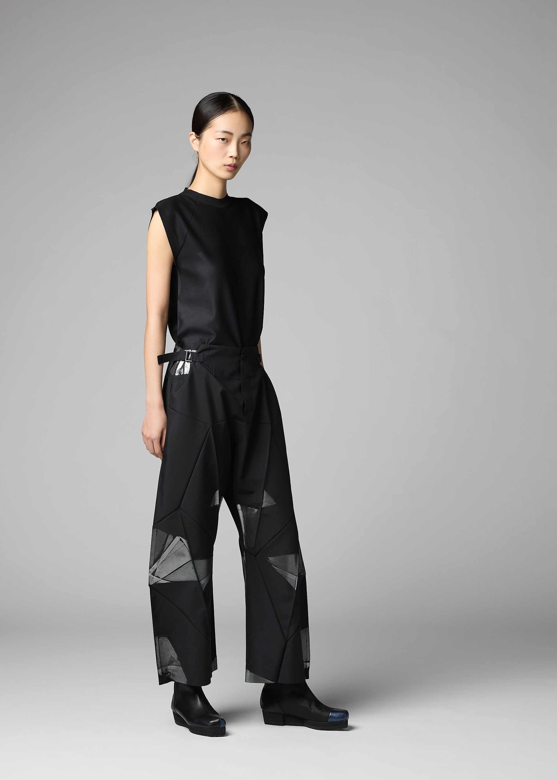 Zoomed view A woman stands in a studio wearing the F-BT from 132 5. ISSEY MIYAKE—sleeveless black top and wide-leg pants with geometric sheer panels, paired with black shoes. She has straight dark hair pulled back, against a plain gray background.