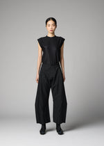 A woman stands against a gray background, wearing the F-BT sleeveless black top and wide adjustable hem pants with geometric seams from 132 5. ISSEY MIYAKE, paired with black boots. Her hair is tied back as she faces the camera neutrally.