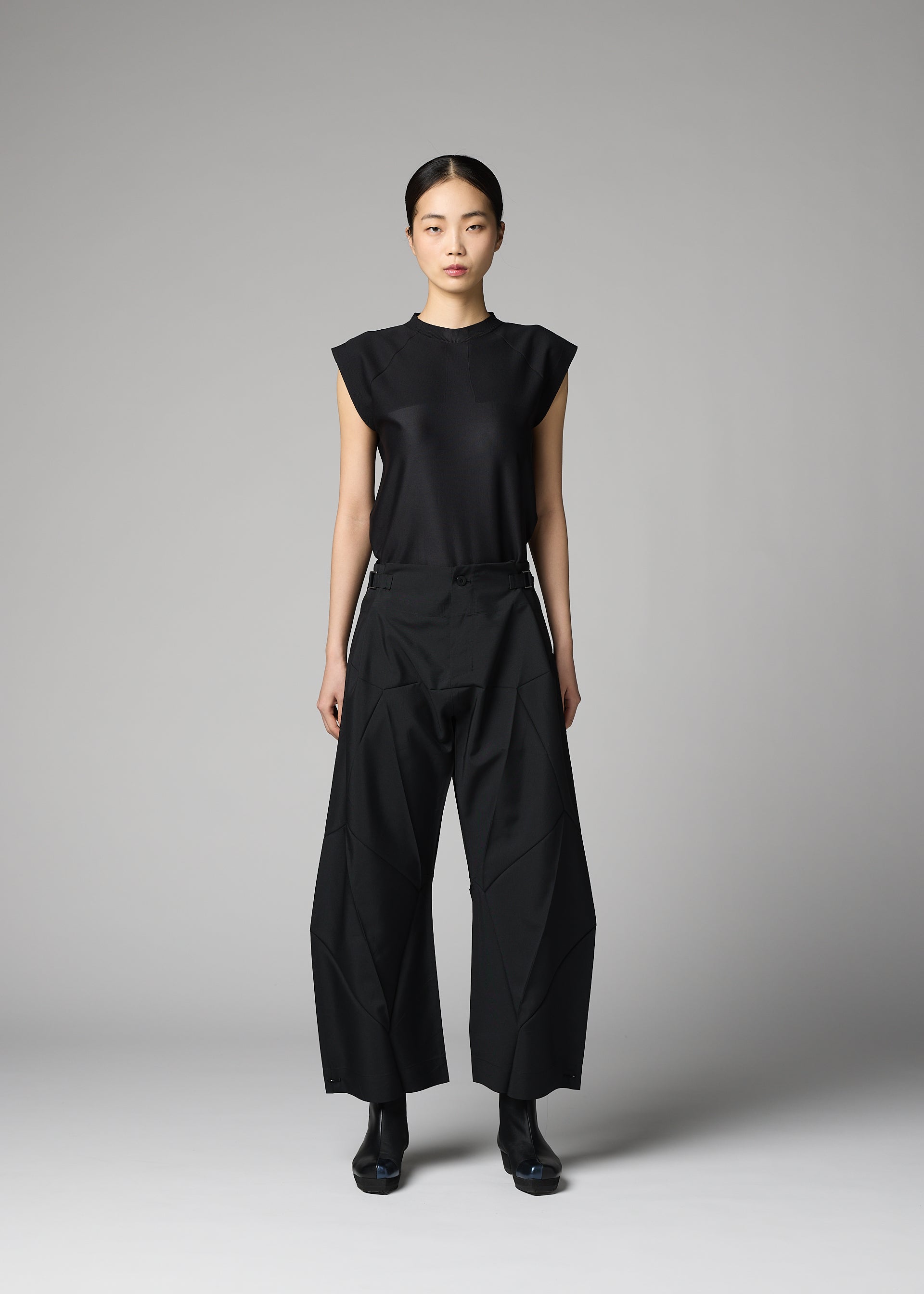 Zoomed view A woman stands against a gray background, wearing the F-BT sleeveless black top and wide adjustable hem pants with geometric seams from 132 5. ISSEY MIYAKE, paired with black boots. Her hair is tied back as she faces the camera neutrally.