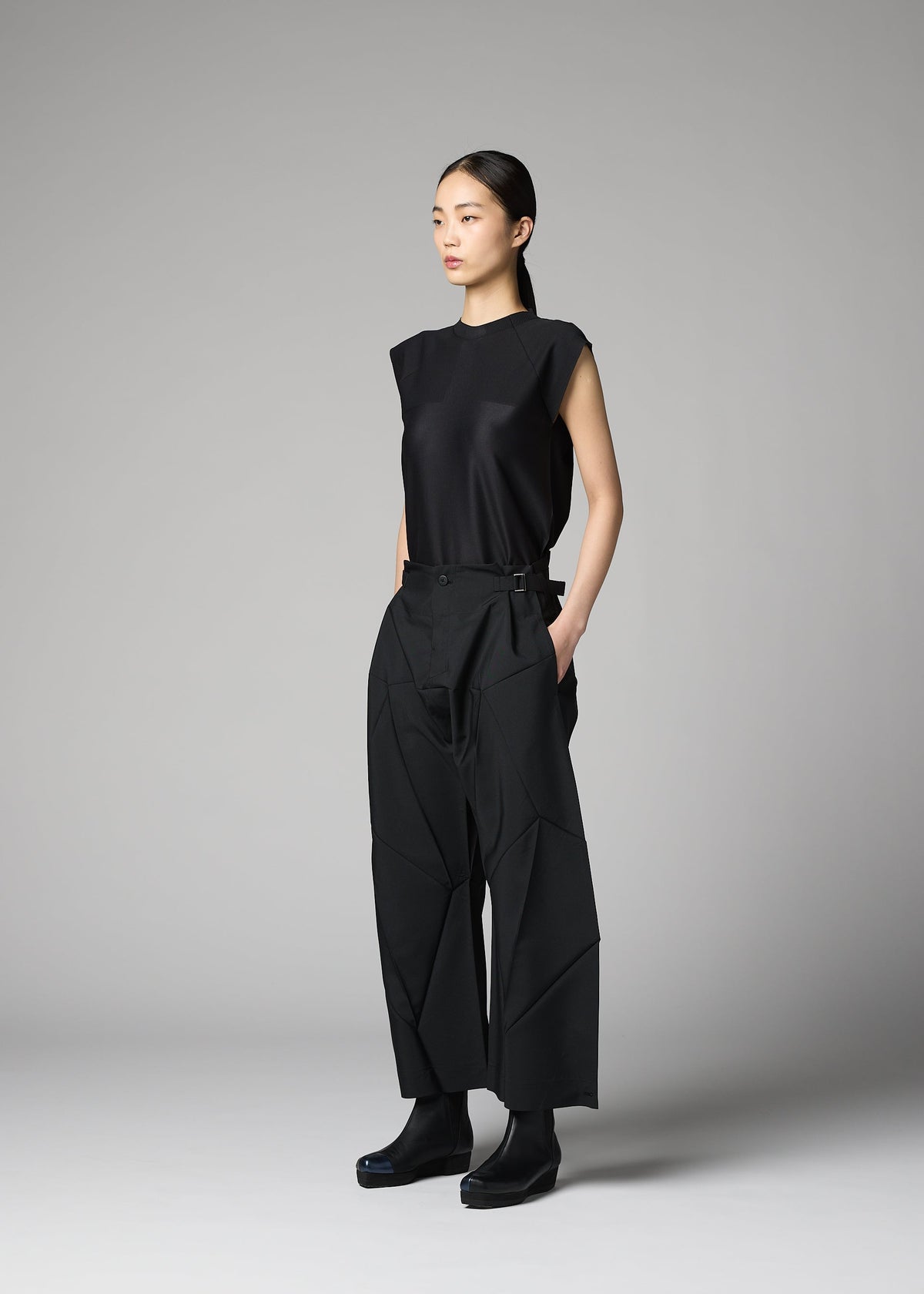 A woman stands in a studio against a plain gray background, wearing the F-BT folding technique pants with geometric seams and a sleeveless black top by 132 5. ISSEY MIYAKE. Her straight dark hair is pulled back and her hands are in her pockets. view 9 of 14