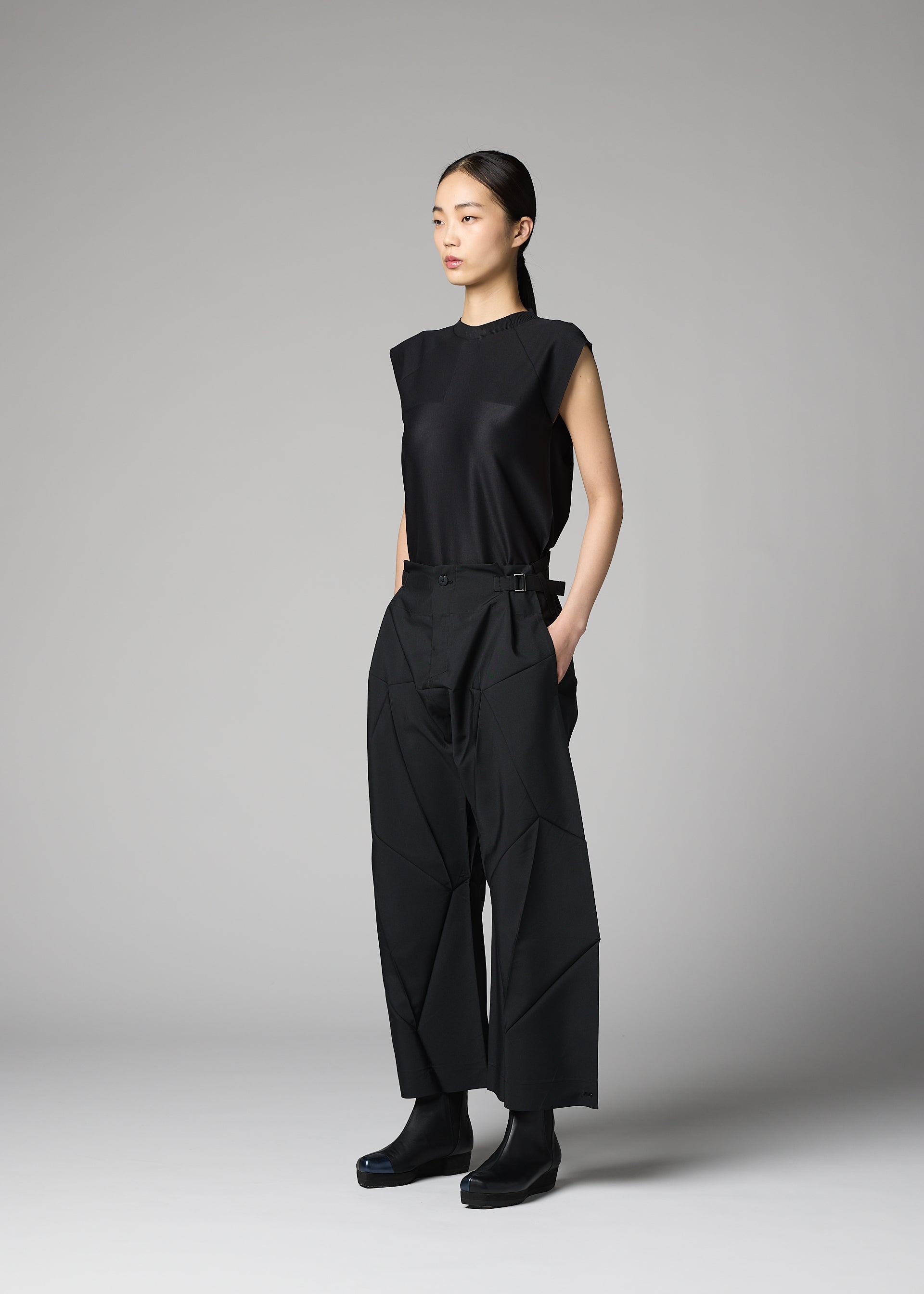 Zoomed view A woman stands in a studio against a plain gray background, wearing the F-BT folding technique pants with geometric seams and a sleeveless black top by 132 5. ISSEY MIYAKE. Her straight dark hair is pulled back and her hands are in her pockets.