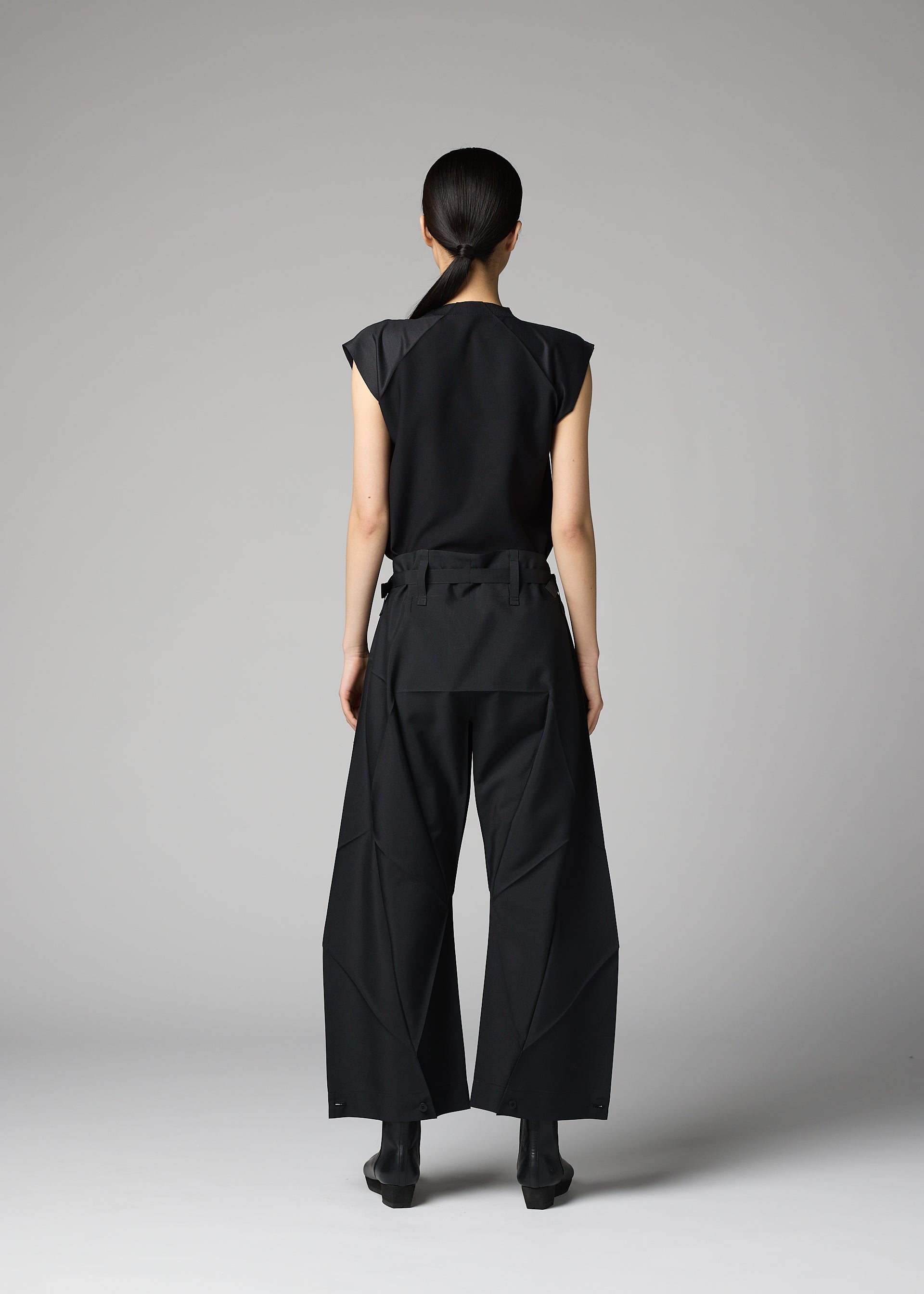 Zoomed view A person with long dark hair, seen from behind, wears the 132 5. ISSEY MIYAKE F-BT black sleeveless top and wide-leg recycled polyester pants with angular seams, paired with black shoes, against a plain light gray background.