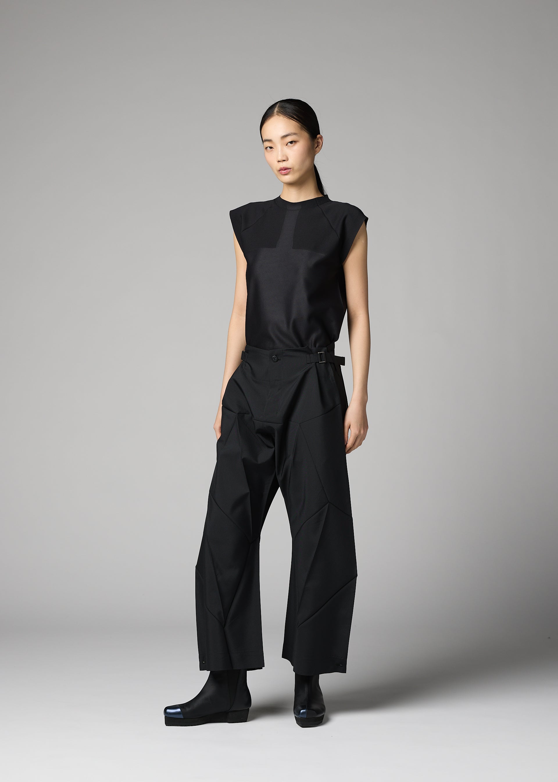 Zoomed view A woman models the F-BT by 132 5. ISSEY MIYAKE—sleeveless black top and belted wide-leg recycled polyester pants—against a grey background, paired with black ankle boots, arms relaxed and expression neutral.