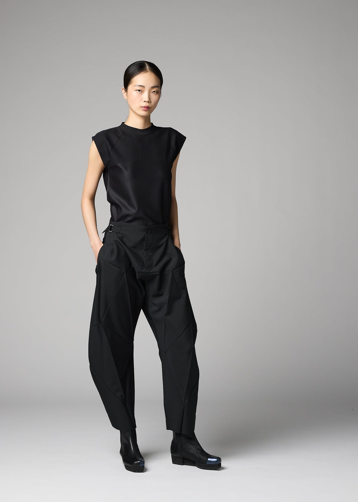 Against a plain gray background, a woman wears the 132 5. ISSEY MIYAKE F-BT sleeveless black top and geometric folding pants, styled with black ankle boots. Her straight hair is pulled back and her hands are in her pockets. view 12 of 14
