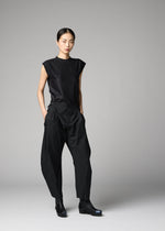 Against a plain gray background, a woman wears the 132 5. ISSEY MIYAKE F-BT sleeveless black top and geometric folding pants, styled with black ankle boots. Her straight hair is pulled back and her hands are in her pockets.