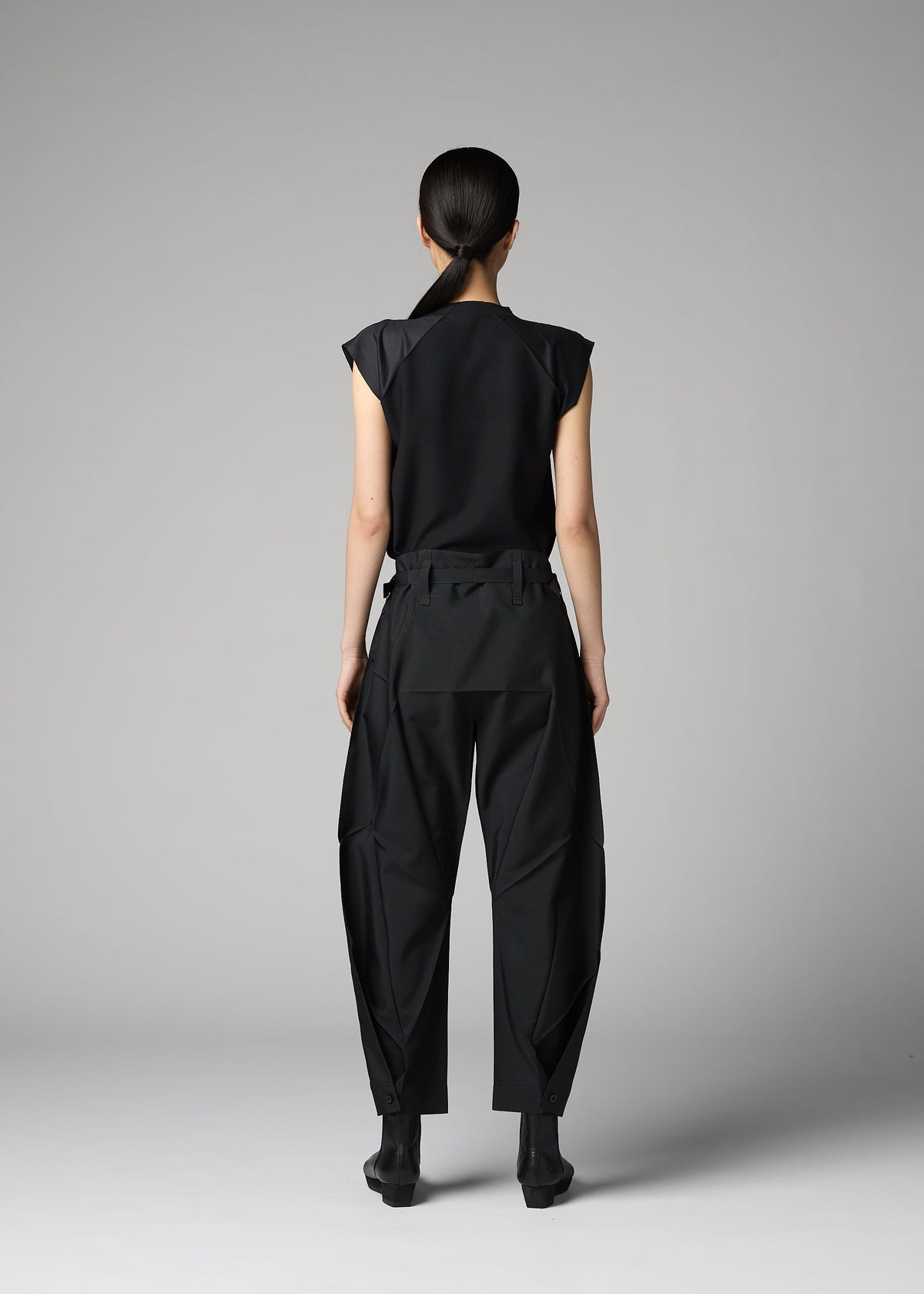 A person stands against a gray background, wearing the F-BT by 132 5. ISSEY MIYAKE—black short-sleeve top, recycled polyester pants with unique seams, and black ankle boots. Their hair is pulled back. view 13 of 14