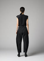 A person stands against a gray background, wearing the F-BT by 132 5. ISSEY MIYAKE—black short-sleeve top, recycled polyester pants with unique seams, and black ankle boots. Their hair is pulled back.