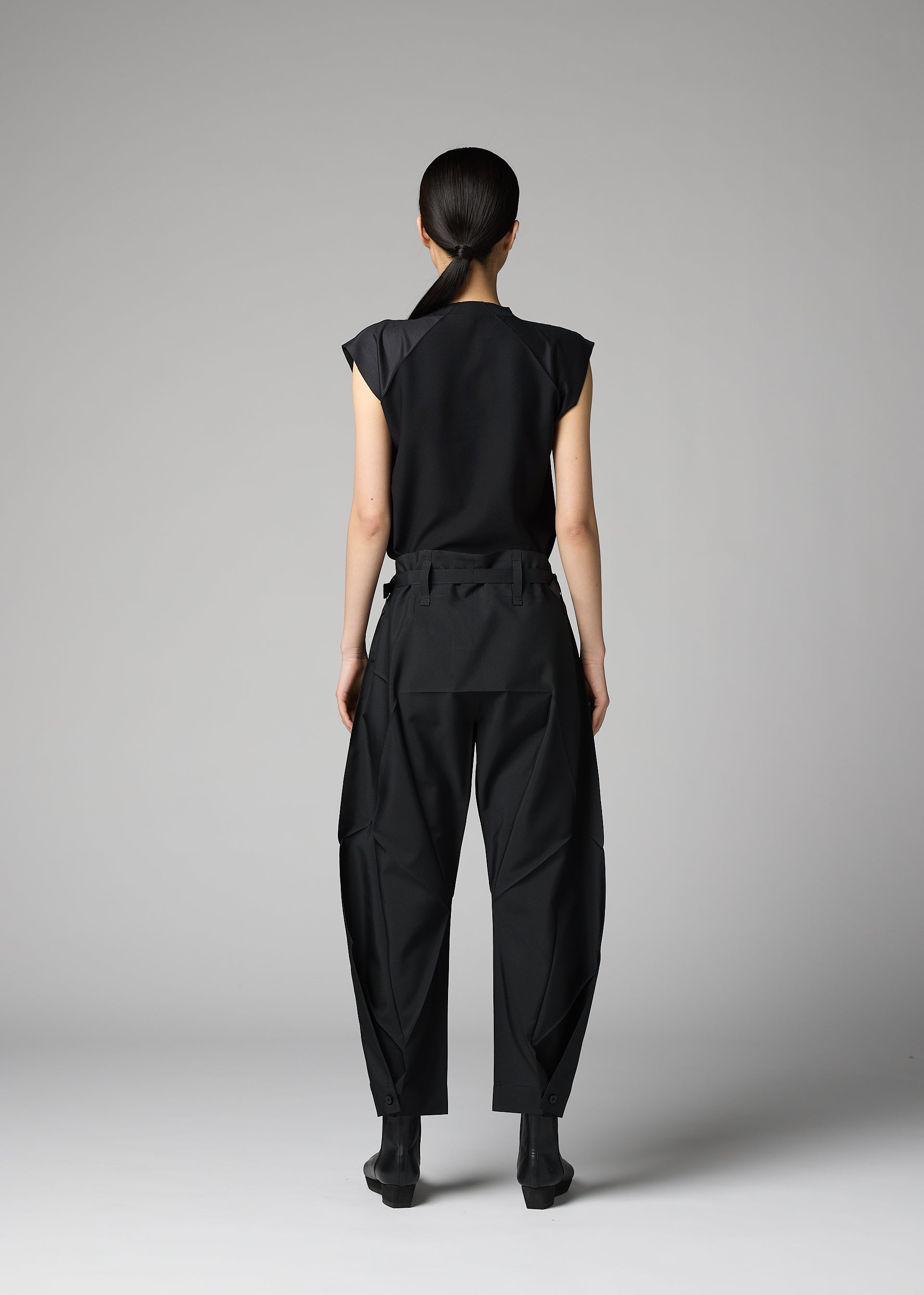 Zoomed view A person stands against a gray background, wearing the F-BT by 132 5. ISSEY MIYAKE—black short-sleeve top, recycled polyester pants with unique seams, and black ankle boots. Their hair is pulled back.