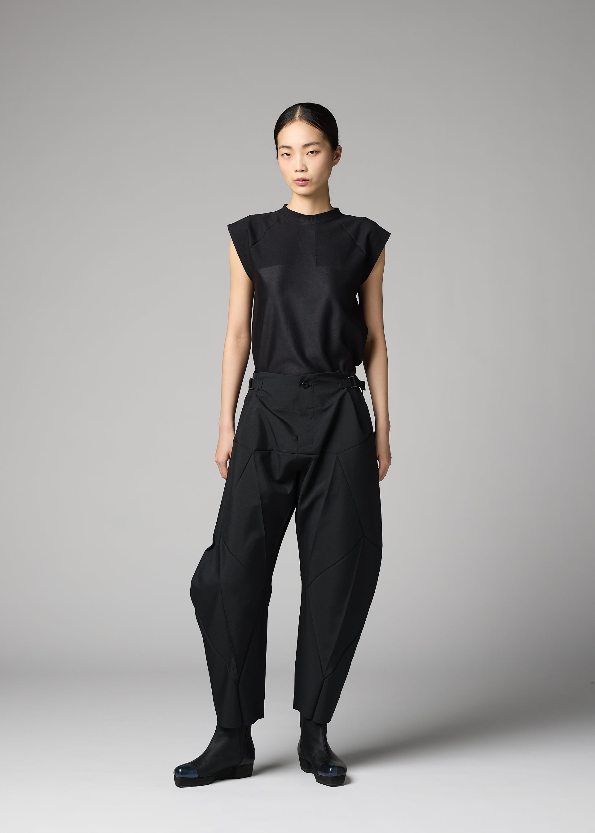 Zoomed view A woman with her hair pulled back stands against a plain gray background, wearing the sleeveless black F-BT top and wide-legged recycled polyester pants with geometric seams by 132 5. ISSEY MIYAKE, paired with black shoes.