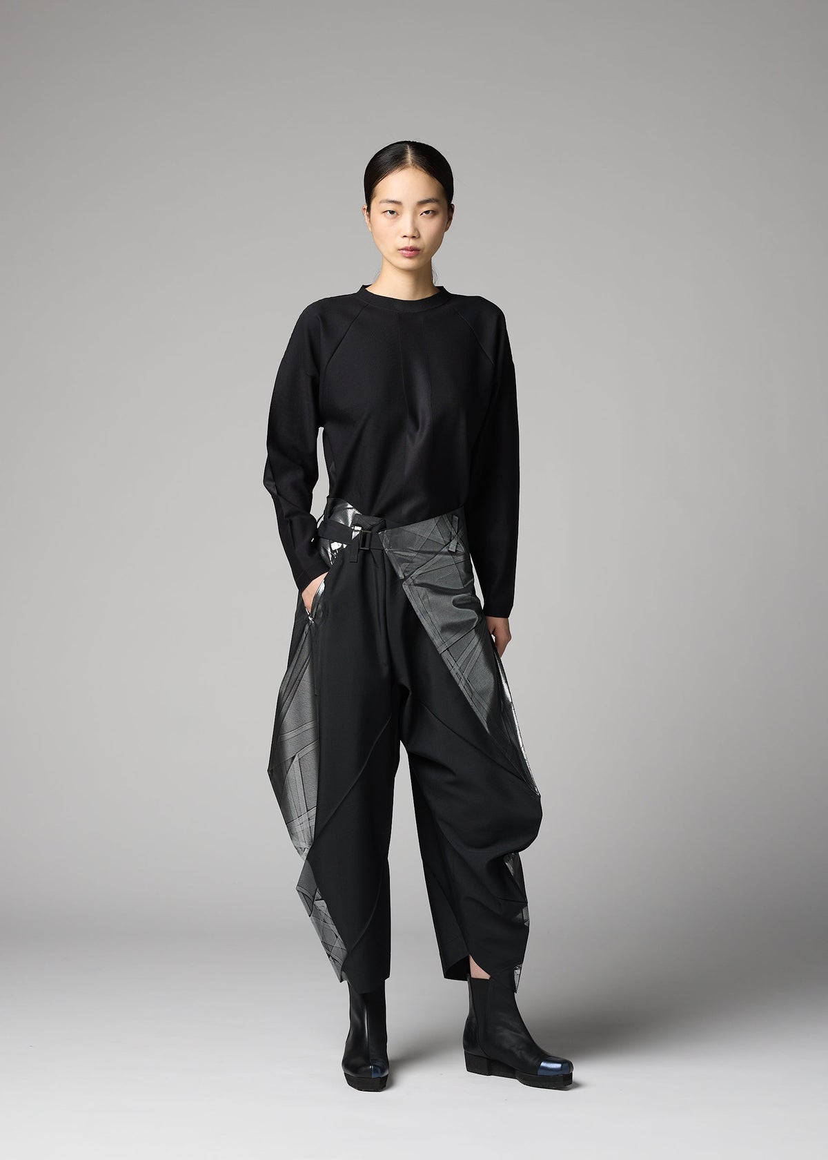 A woman faces forward with a neutral expression, wearing the F-BT black long-sleeve top and shiny gray foil-processed black recycled polyester pants by 132 5. ISSEY MIYAKE. Her hair is tied back, and she completes the look with black boots against a gray background. view 6 of 9
