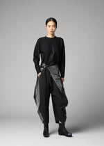 A woman faces forward with a neutral expression, wearing the F-BT black long-sleeve top and shiny gray foil-processed black recycled polyester pants by 132 5. ISSEY MIYAKE. Her hair is tied back, and she completes the look with black boots against a gray background.