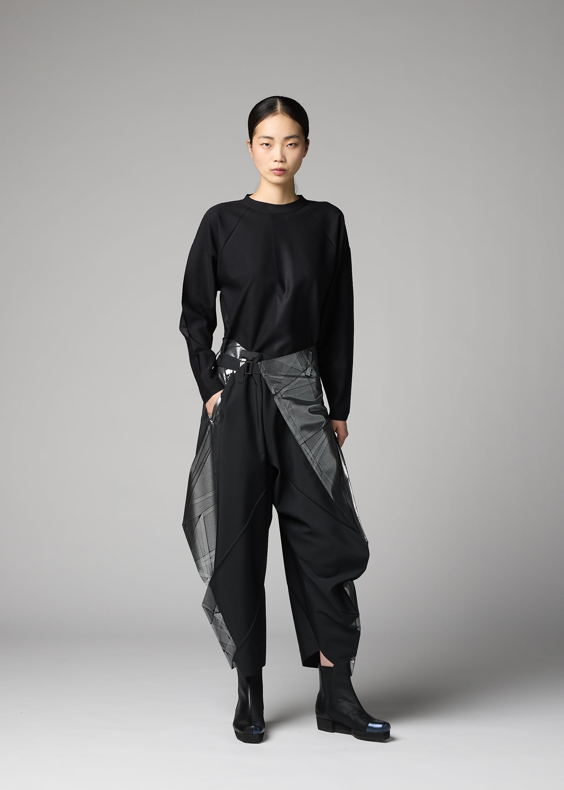 Zoomed view A woman faces forward with a neutral expression, wearing the F-BT black long-sleeve top and shiny gray foil-processed black recycled polyester pants by 132 5. ISSEY MIYAKE. Her hair is tied back, and she completes the look with black boots against a gray background.