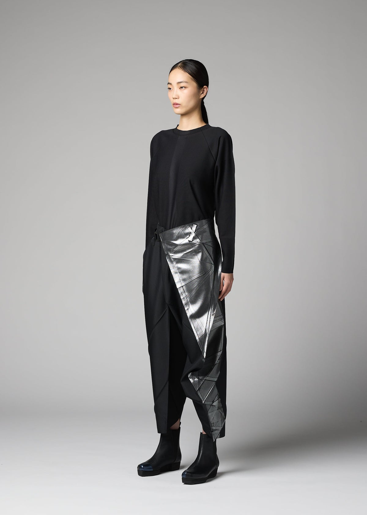 A woman with long dark hair tied back stands against a gray background, wearing the F-BT metallic silver asymmetrical wrap skirt by 132 5. ISSEY MIYAKE, paired with a black top, recycled polyester pants, and black ankle boots. view 7 of 9