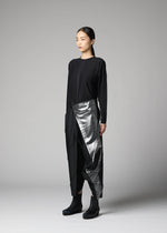 A woman with long dark hair tied back stands against a gray background, wearing the F-BT metallic silver asymmetrical wrap skirt by 132 5. ISSEY MIYAKE, paired with a black top, recycled polyester pants, and black ankle boots.