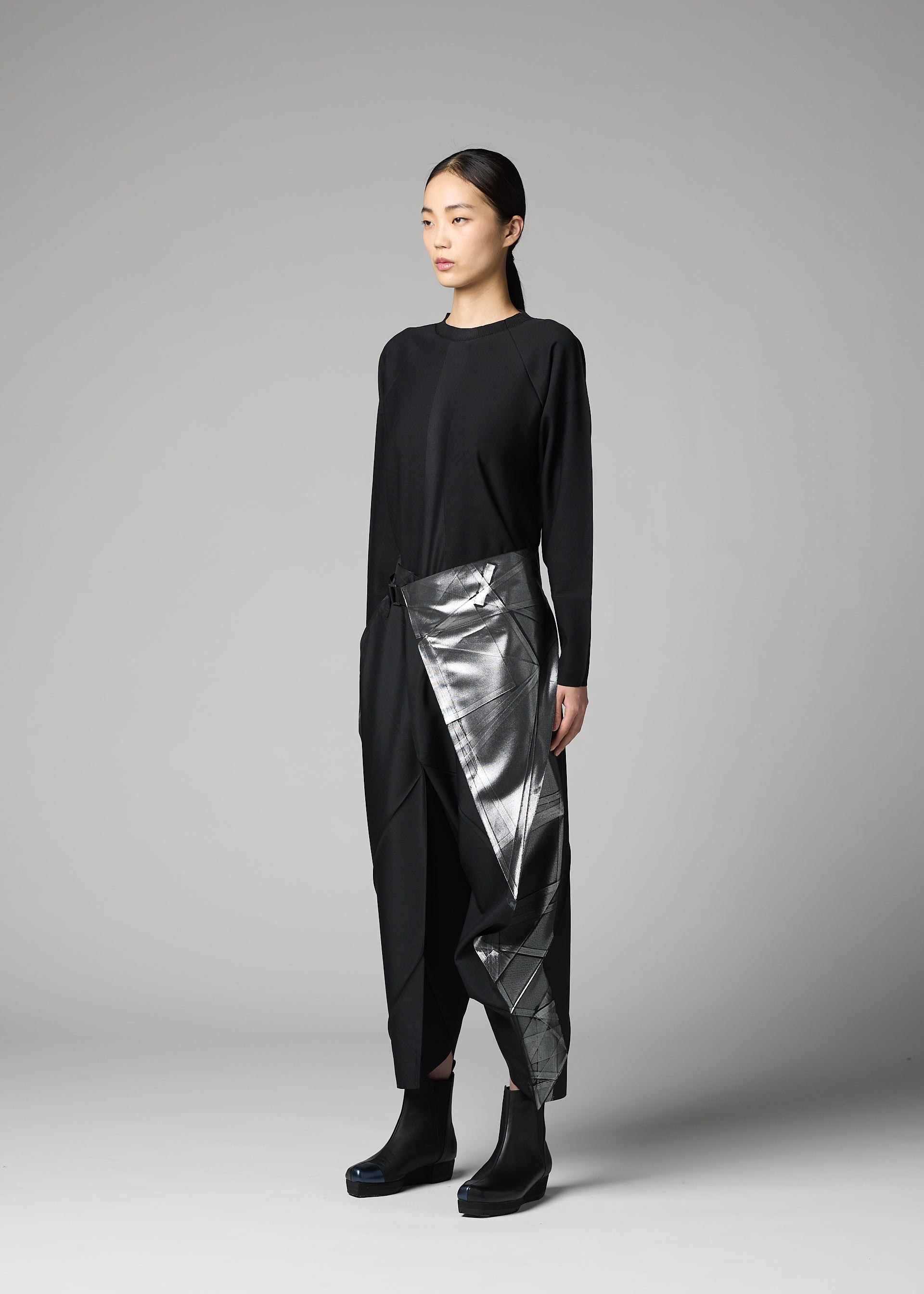 Zoomed view A woman with long dark hair tied back stands against a gray background, wearing the F-BT metallic silver asymmetrical wrap skirt by 132 5. ISSEY MIYAKE, paired with a black top, recycled polyester pants, and black ankle boots.