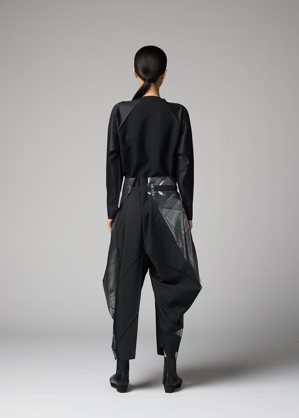 In a minimalist studio, a person with tied-back hair models the F-BT pants by 132 5. ISSEY MIYAKE—black recycled polyester trousers with geometric sheer-panel details—paired with a black long-sleeve top and black ankle boots, facing away. view 8 of 9