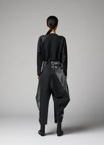 In a minimalist studio, a person with tied-back hair models the F-BT pants by 132 5. ISSEY MIYAKE—black recycled polyester trousers with geometric sheer-panel details—paired with a black long-sleeve top and black ankle boots, facing away.