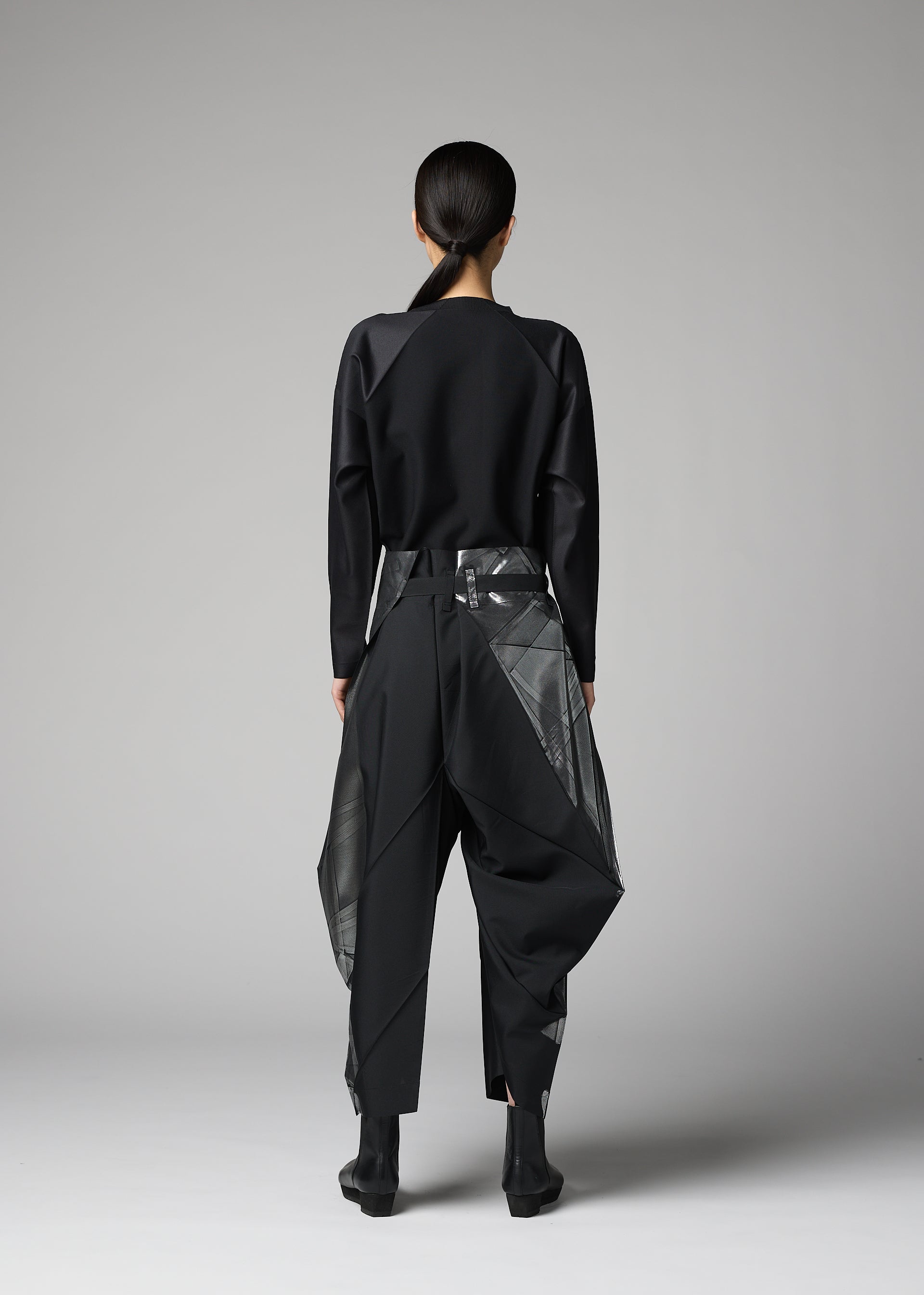 Zoomed view In a minimalist studio, a person with tied-back hair models the F-BT pants by 132 5. ISSEY MIYAKE—black recycled polyester trousers with geometric sheer-panel details—paired with a black long-sleeve top and black ankle boots, facing away.