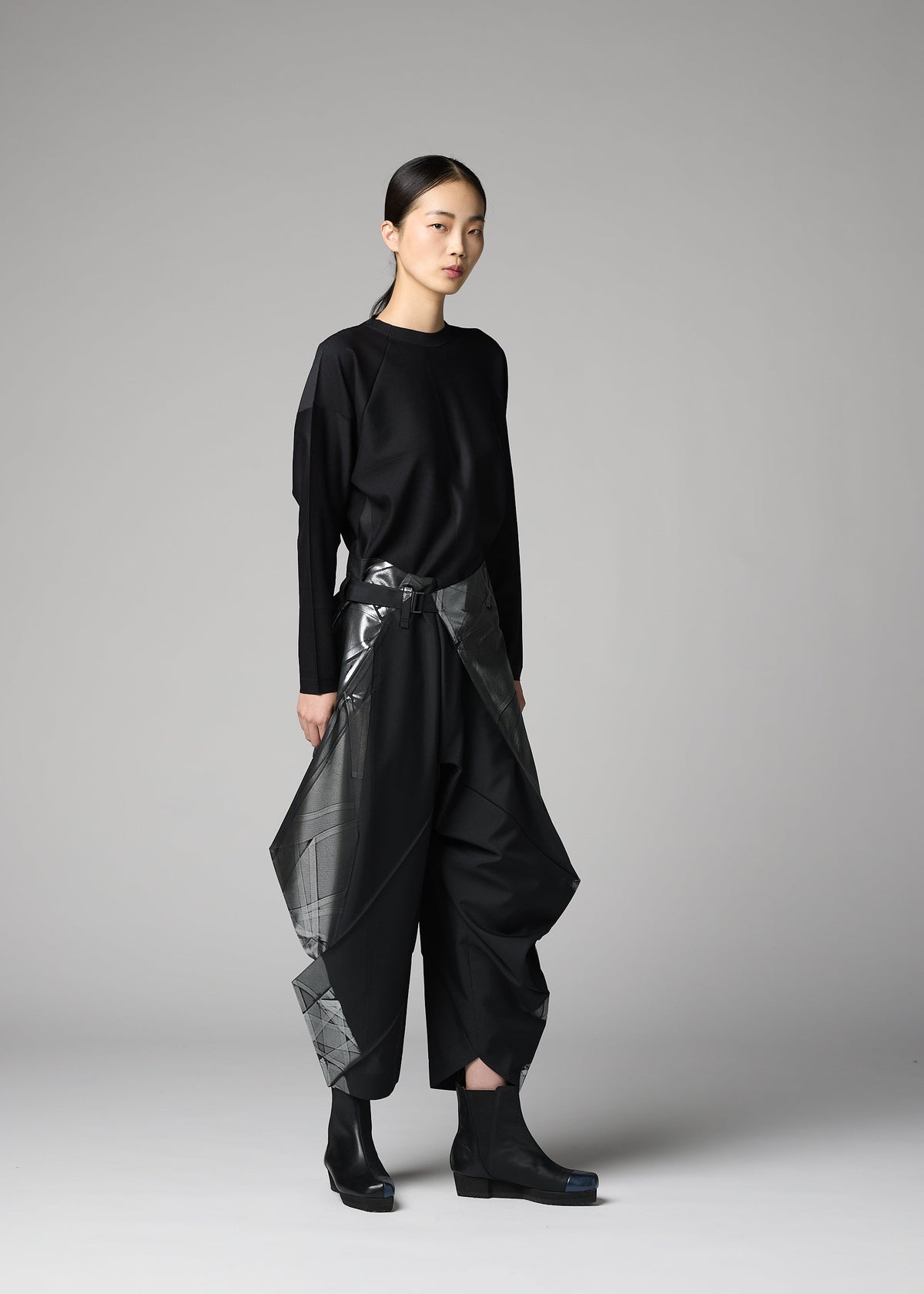 A person stands against a plain gray background, wearing the F-BT by 132 5. ISSEY MIYAKE: a loose black long-sleeve top and wide recycled polyester pants with shiny angular panels, paired with black boots. Their hair is tied back and expression is neutral. view 9 of 9