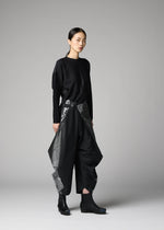 A person stands against a plain gray background, wearing the F-BT by 132 5. ISSEY MIYAKE: a loose black long-sleeve top and wide recycled polyester pants with shiny angular panels, paired with black boots. Their hair is tied back and expression is neutral.