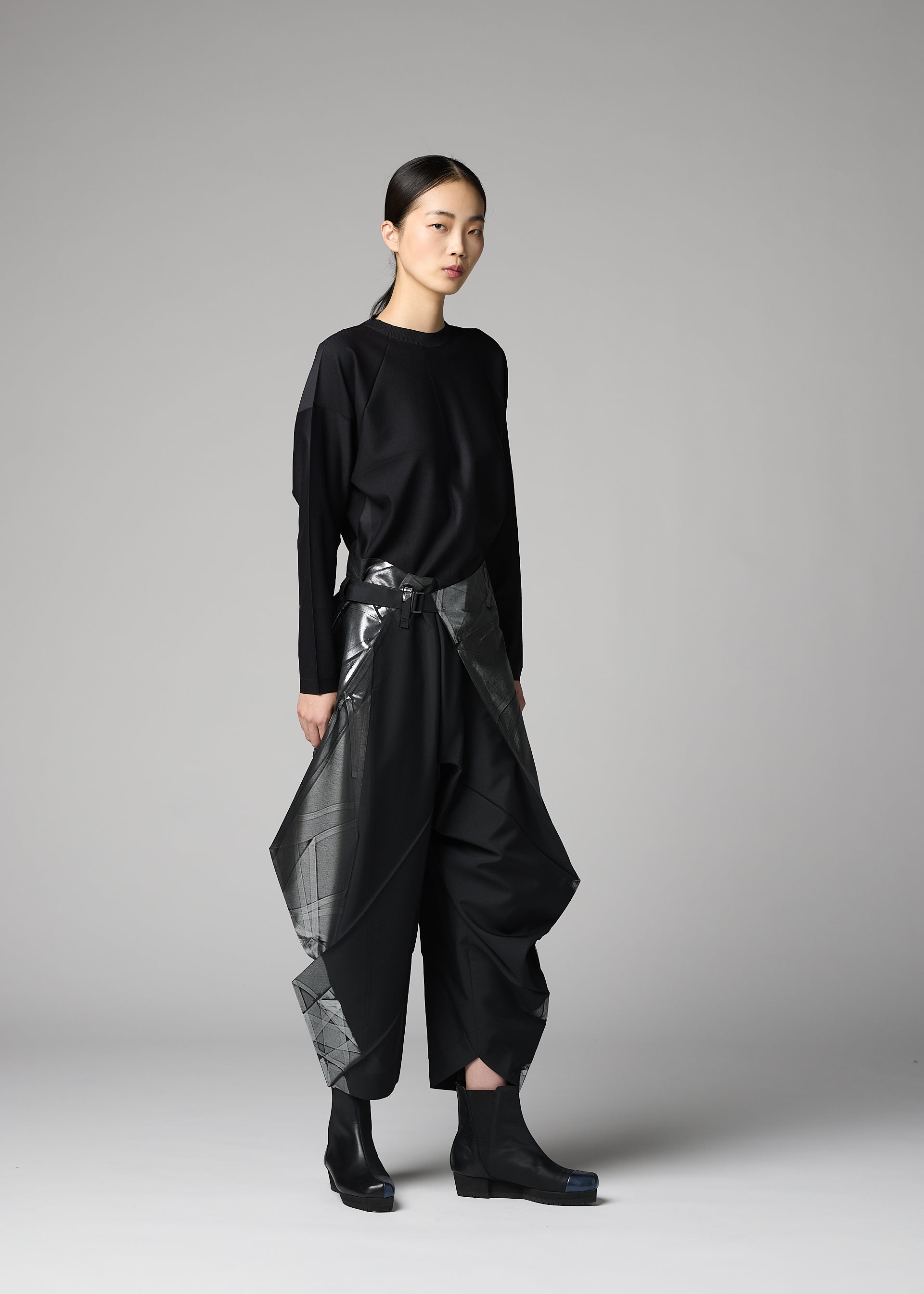 Zoomed view A person stands against a plain gray background, wearing the F-BT by 132 5. ISSEY MIYAKE: a loose black long-sleeve top and wide recycled polyester pants with shiny angular panels, paired with black boots. Their hair is tied back and expression is neutral.