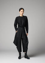A woman stands in a studio wearing the F-BT pants by 132 5. ISSEY MIYAKE, styled with a black long-sleeve top and ankle boots. A plain gray backdrop completes the minimalist look.