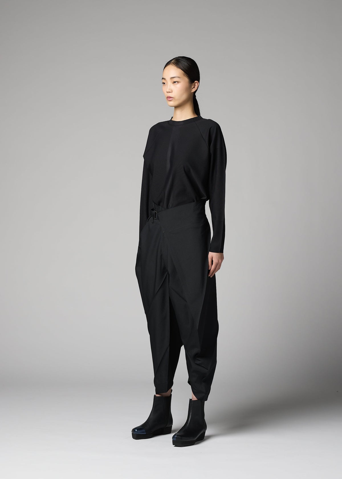 A person stands against a gray background, wearing the F-BT pants by 132 5. ISSEY MIYAKE—black folding technique trousers paired with a black long-sleeve top and platform ankle boots. Their straight dark hair is tied back as they look slightly to the side. view 7 of 9