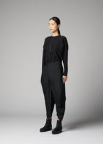 A person stands against a gray background, wearing the F-BT pants by 132 5. ISSEY MIYAKE—black folding technique trousers paired with a black long-sleeve top and platform ankle boots. Their straight dark hair is tied back as they look slightly to the side.